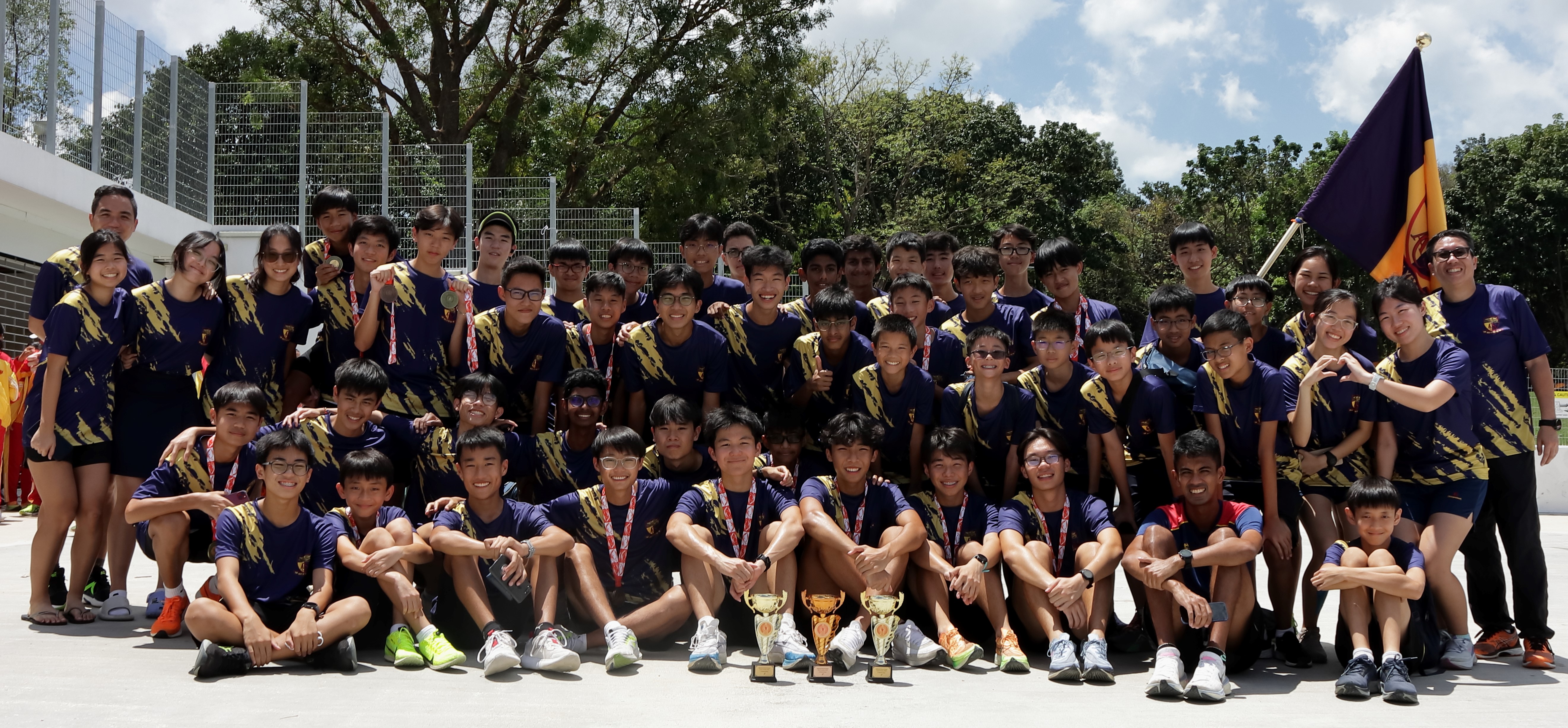 Large group of students in matching navy and gold uniforms posing outdoors with trophies, medals, and a purple flag.